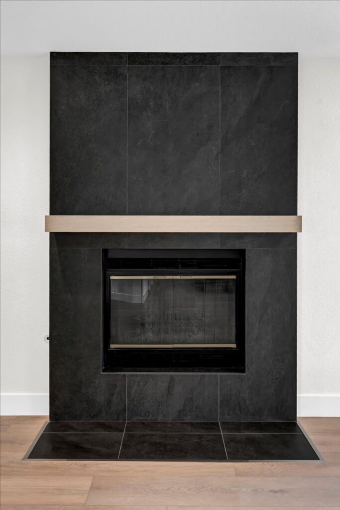 Living room fireplace with charcoal tile surround, light wood mantel, dark hearth and wood flooring.