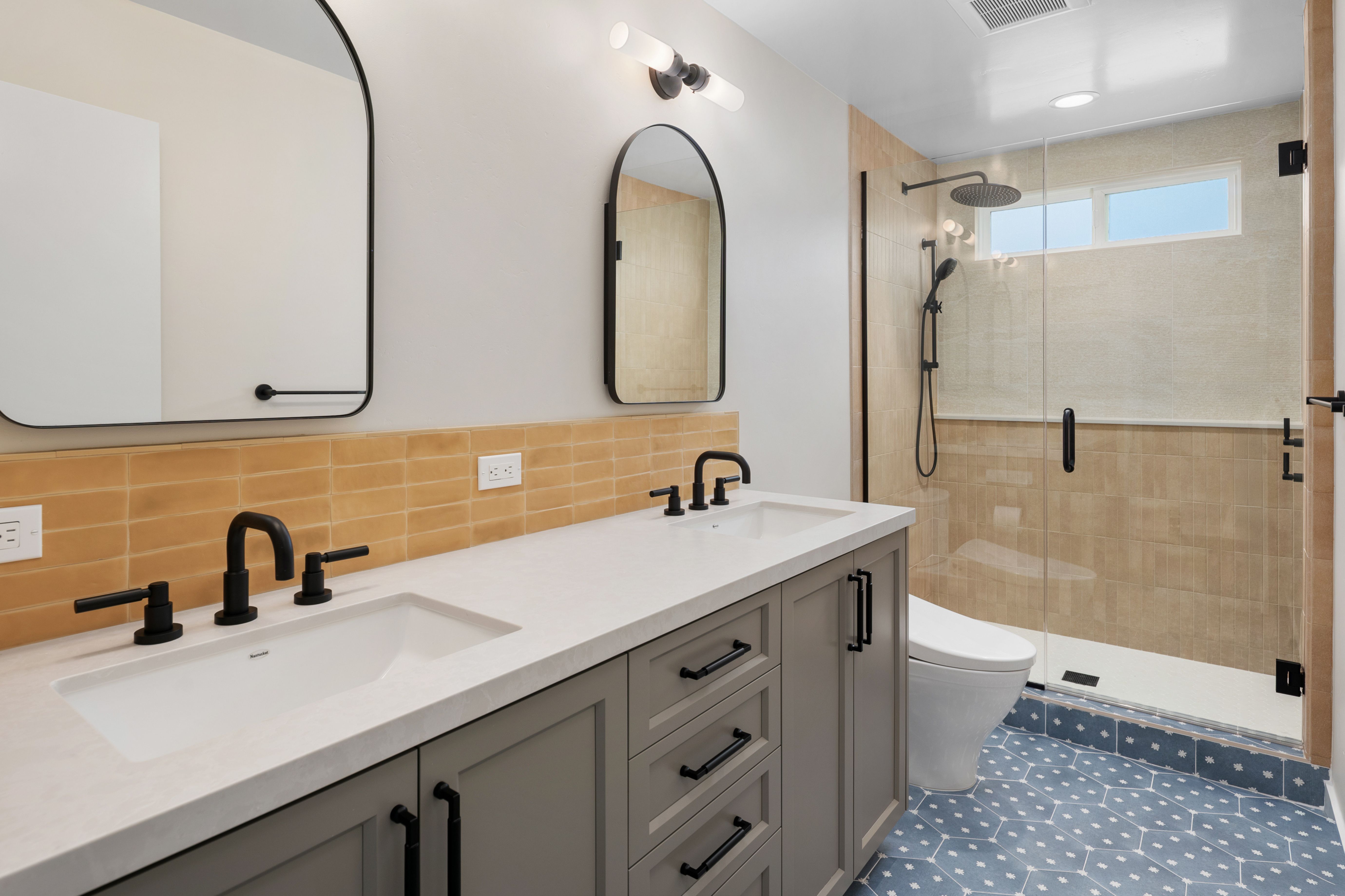Full bathroom with double vanity, black fixtures, amber tile backsplash, glass shower and patterned blue floor tile.