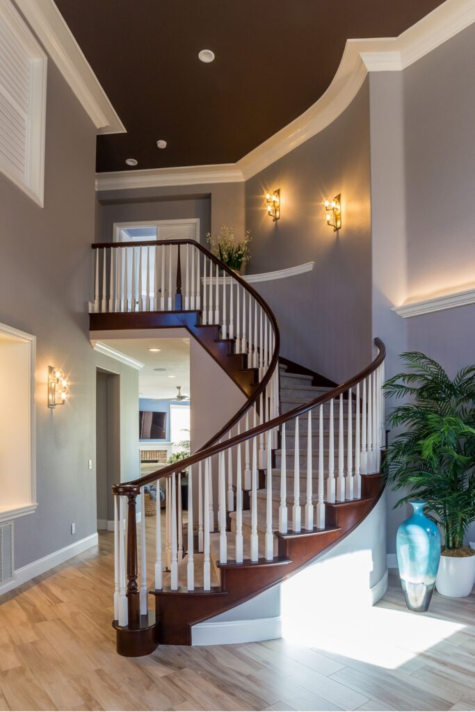 Curved staircase with dark wood handrail, white balusters, gray walls, crown molding and wall sconces.