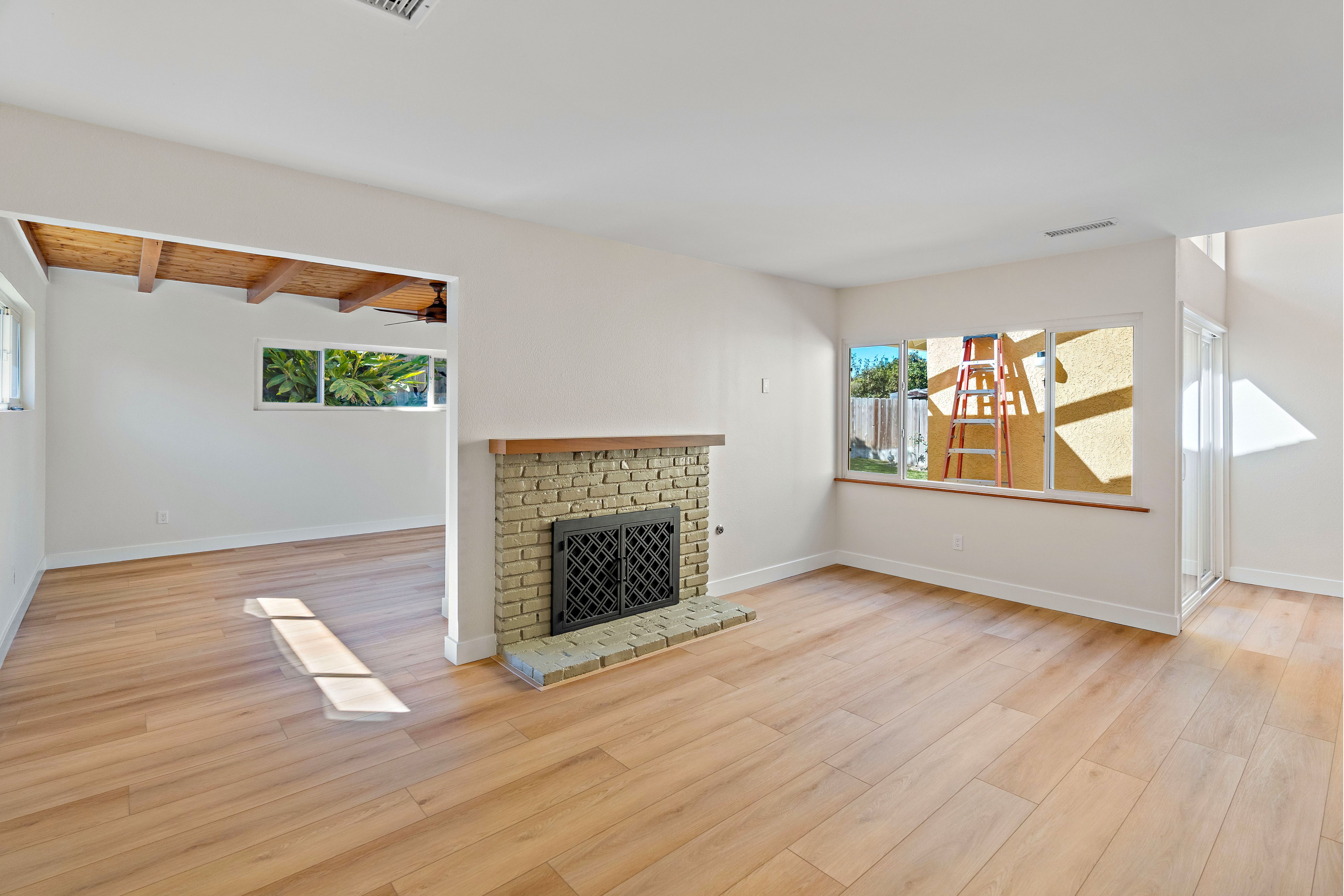 Open living area with brick fireplace, wood mantel, wide-plank flooring, large windows and exposed ceiling beams.