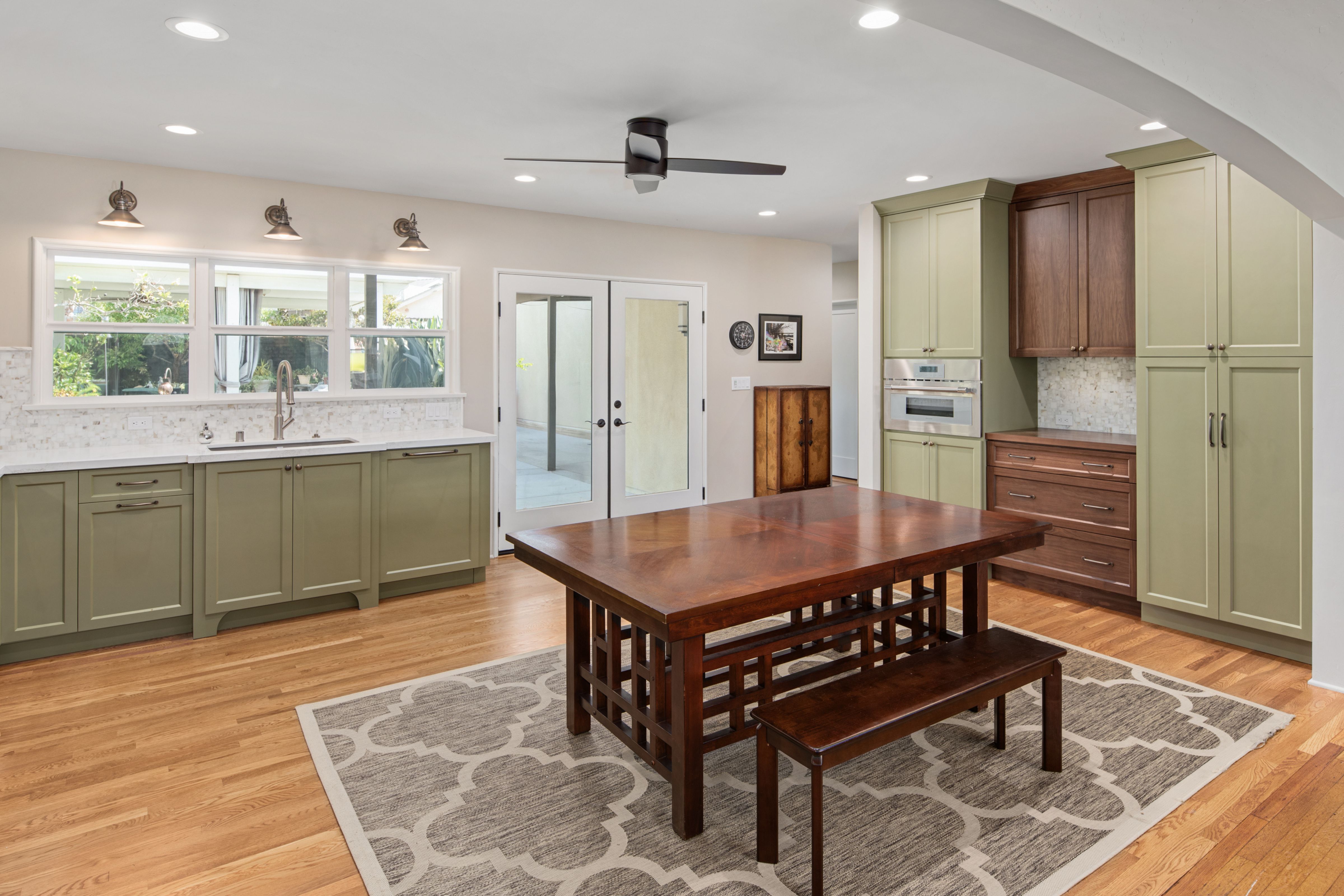 Kitchen with sage cabinetry, wood accents, hardwood floors, dining table, French doors and window-lined sink wall.