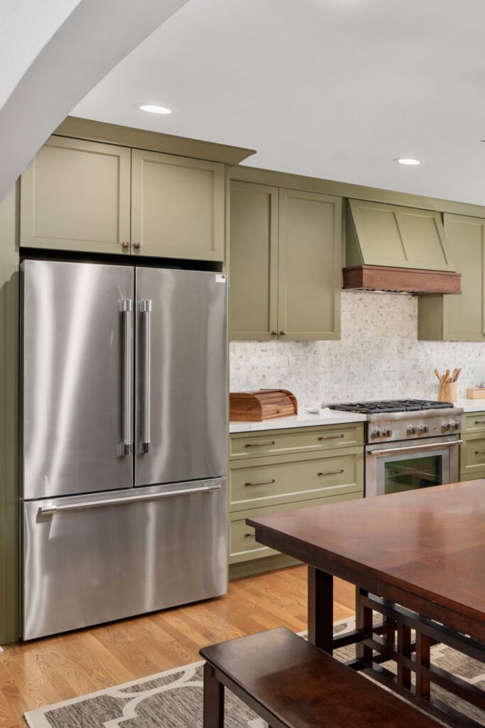 Kitchen range and stainless refrigerator framed by sage cabinetry, mosaic tile, and wood dining furniture.