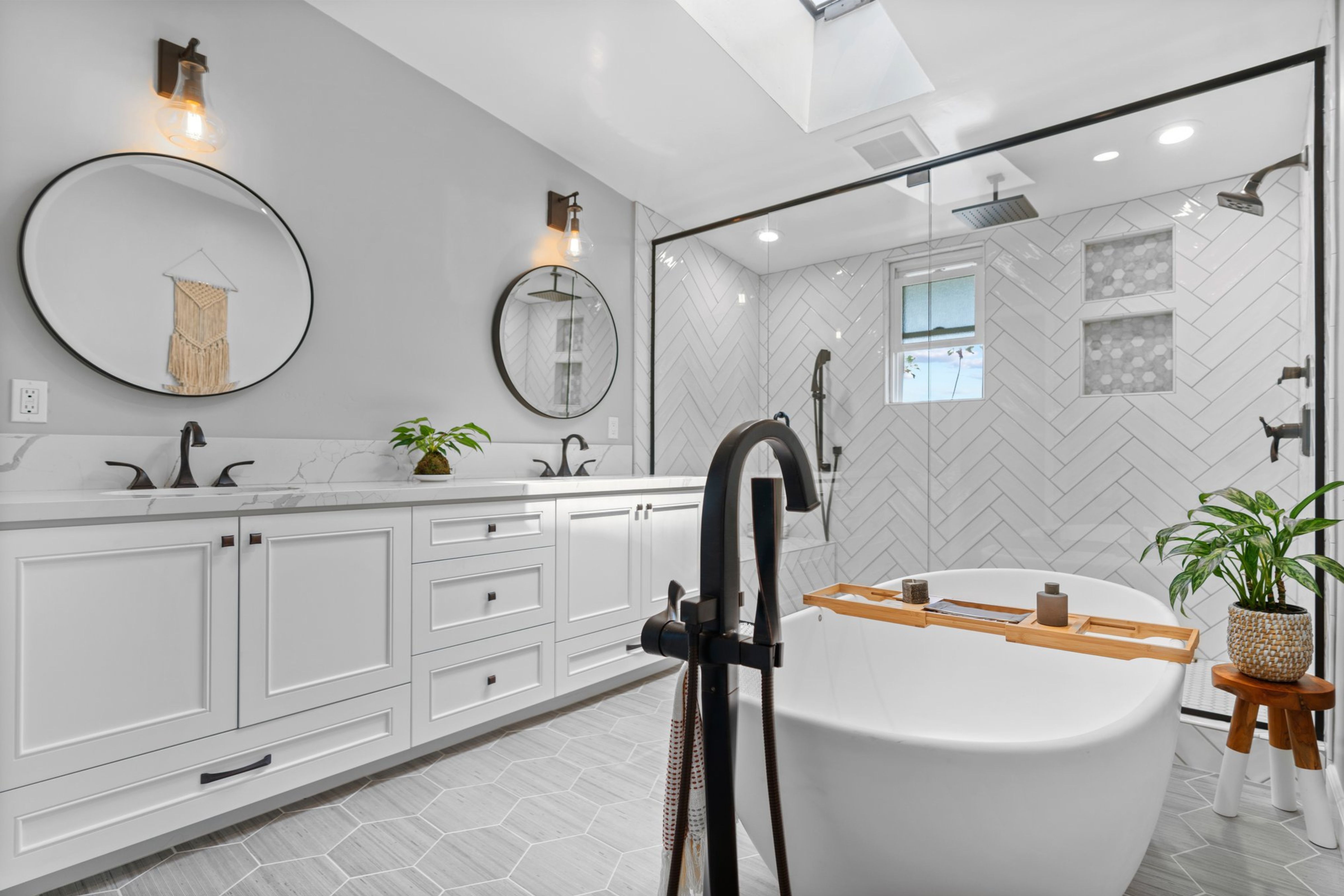 Primary bathroom with white double vanity, freestanding tub, glass shower, herringbone tile and dark bronze fixtures.