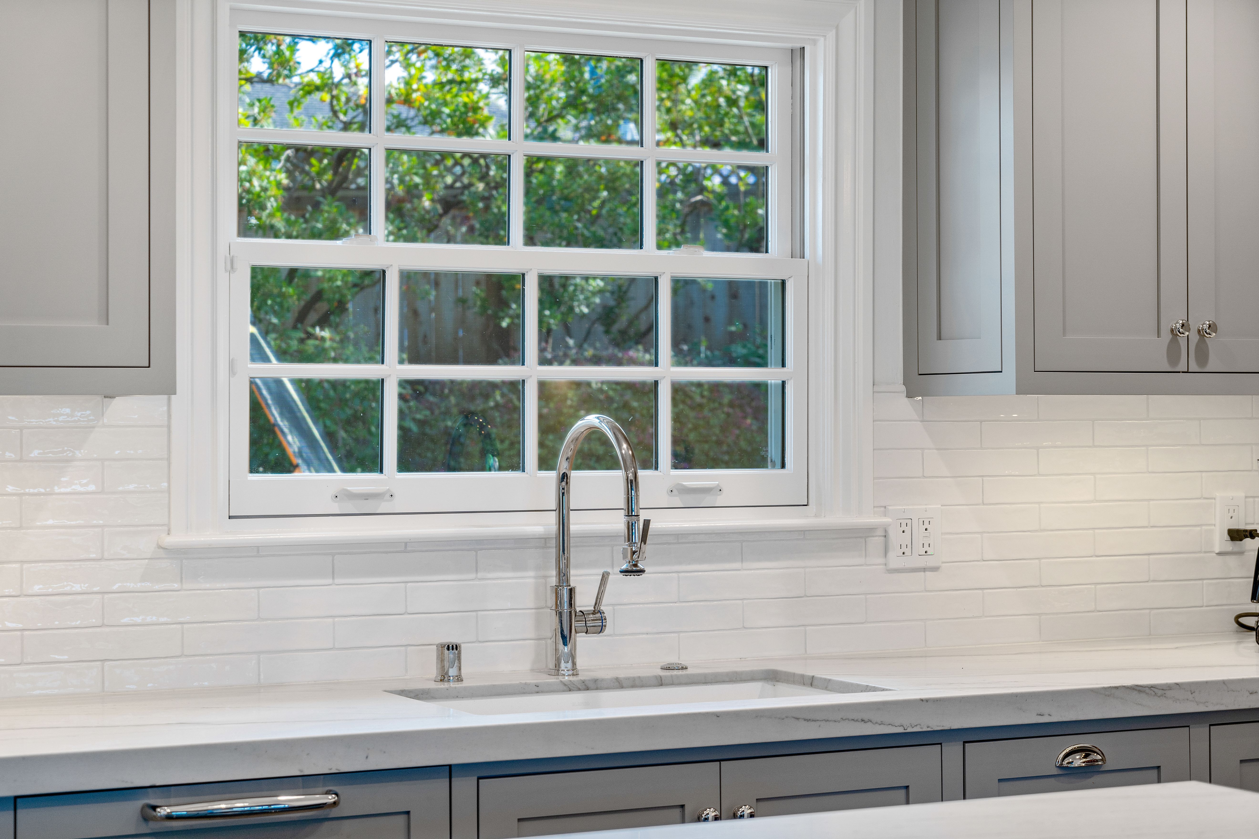 Kitchen sink beneath a divided-light window with painted cabinetry, white tile backsplash and polished faucet.