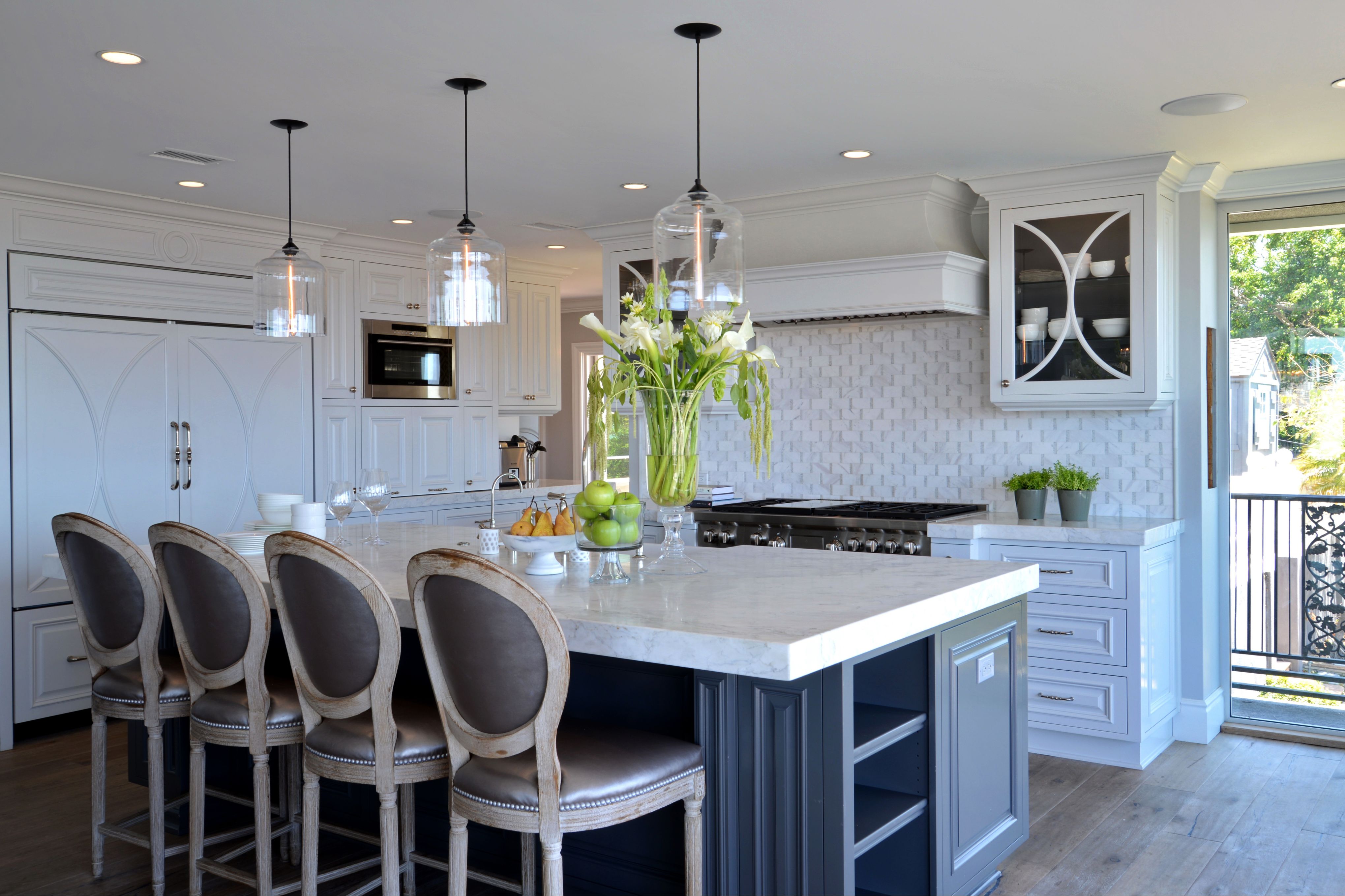 Remodeled kitchen with white cabinetry, dark island, marble counters, pendant lights and decorative range hood.