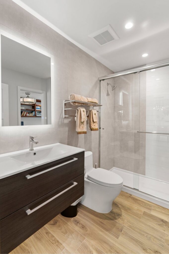 Bathroom with dark floating vanity, illuminated mirror, glass shower, gray wall tile and wood-look flooring.