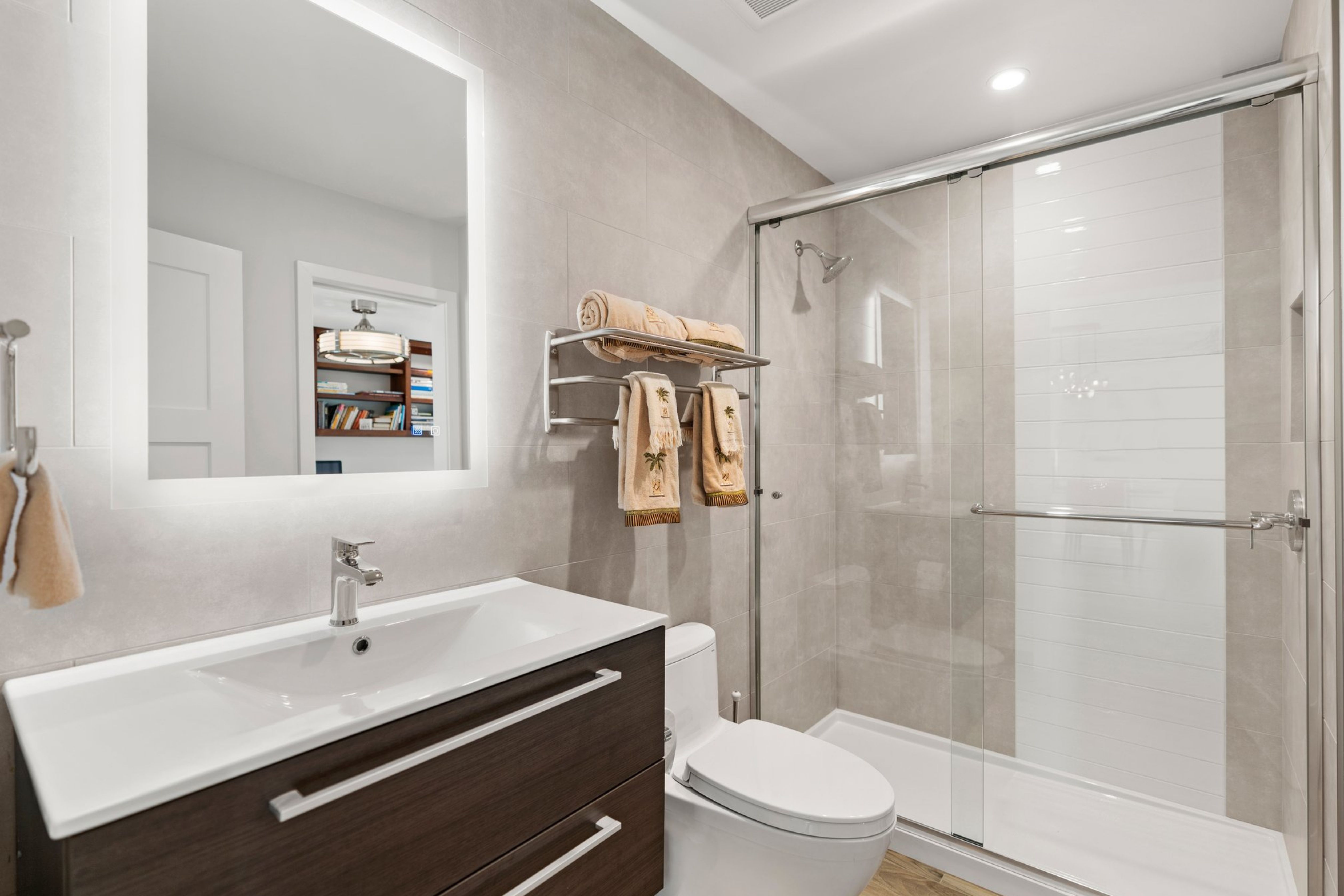 Modern bathroom with glass shower, large-format tile, dark vanity, illuminated mirror and wood-look flooring.