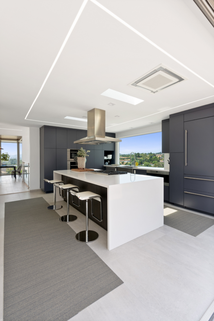 Modern kitchen with dark flat-panel cabinetry, white waterfall island, skylights, linear lighting and expansive windows.