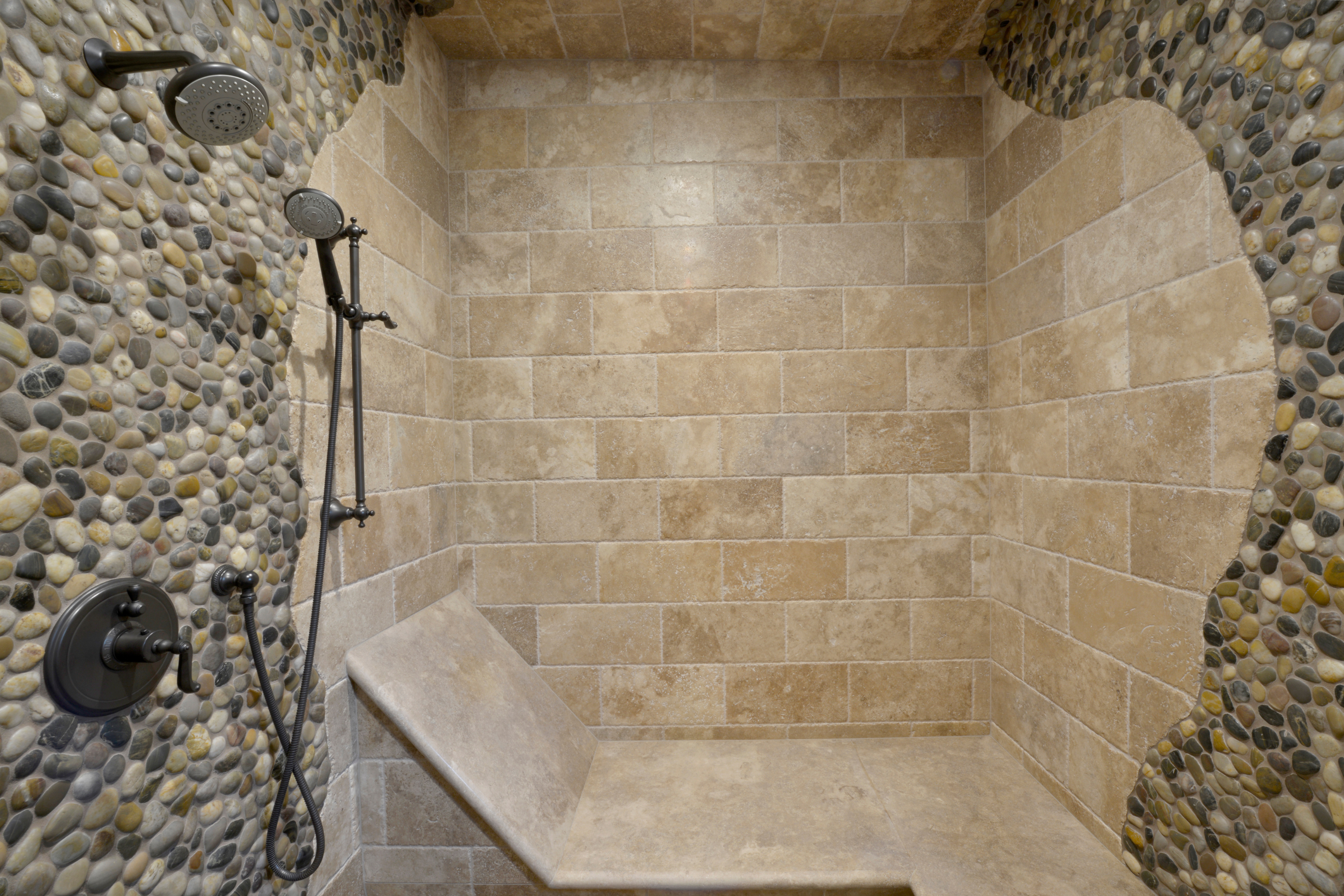 Walk-in shower with stone tile walls, pebble accents, built-in bench and dual shower fixtures.