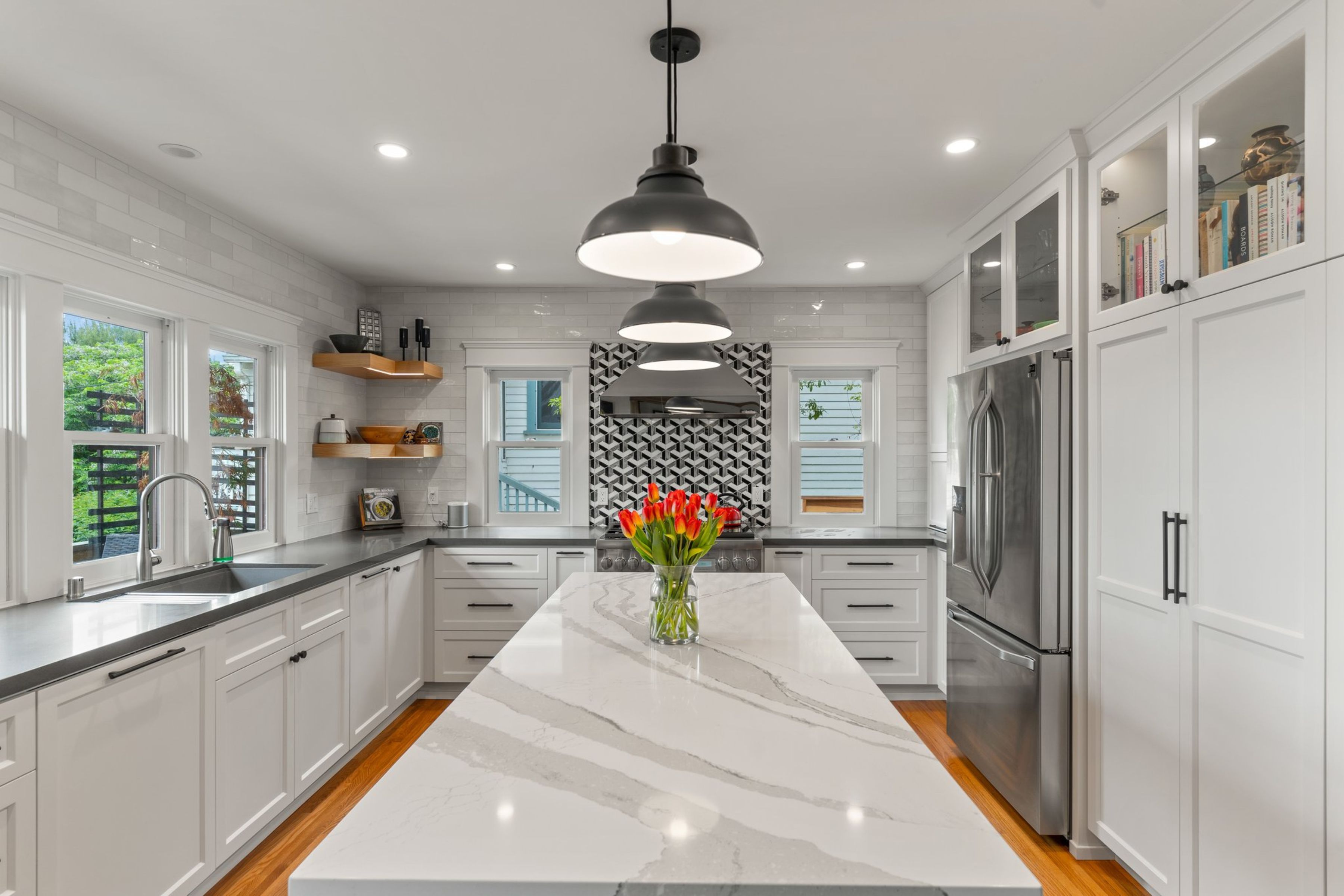 White kitchen with long island, stainless appliances, pendant lighting, geometric range backsplash and hardwood floors.