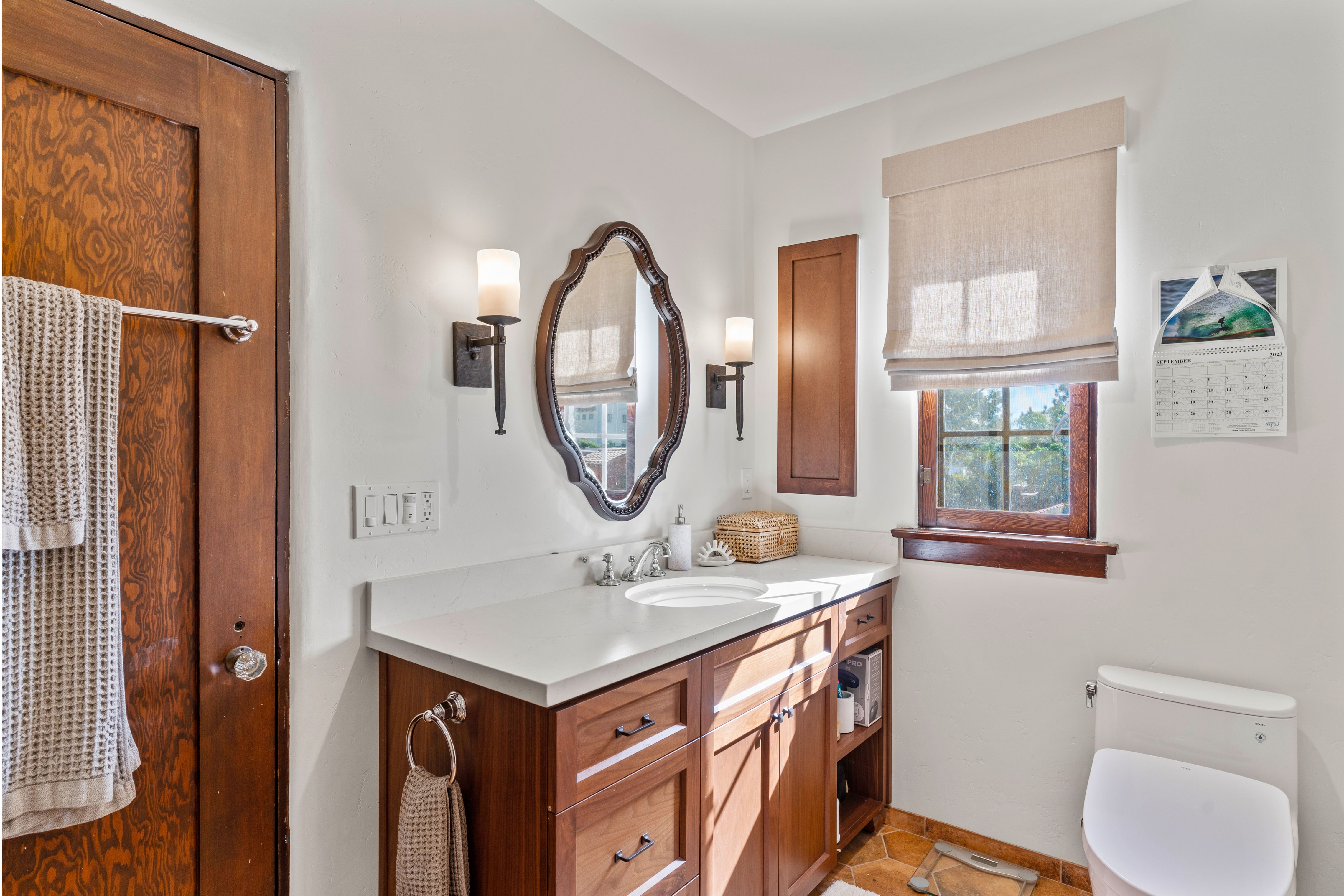 Bathroom with wood vanity, light countertop, framed oval mirror, wall sconces and warm-toned tile flooring.