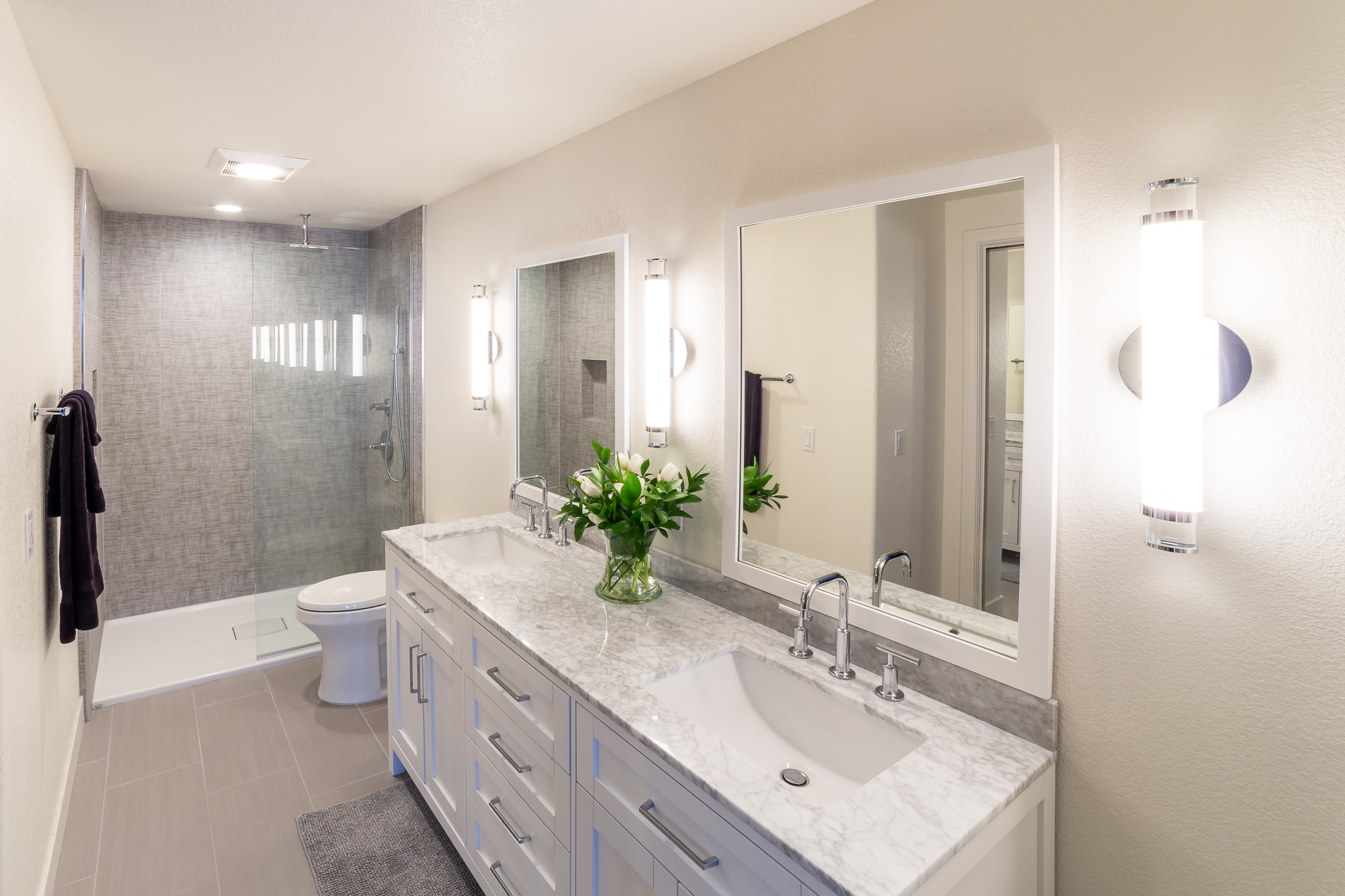 Primary bathroom with long white vanity, stone countertop, frameless glass shower, gray tile and vertical sconces.