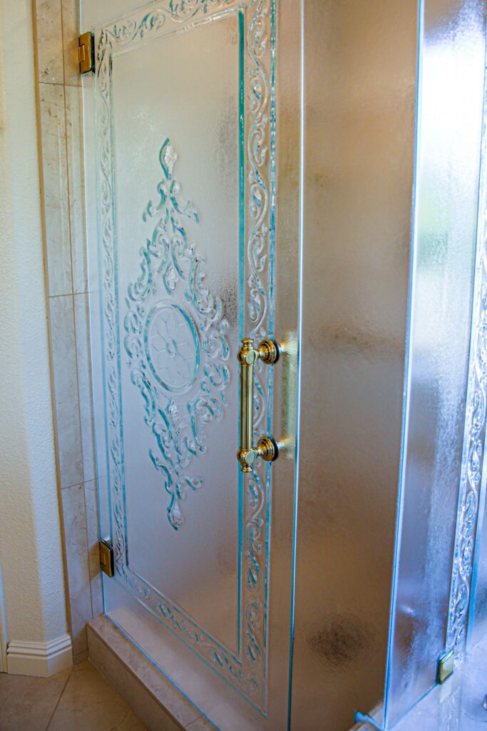Etched glass shower door with ornate scrollwork, frosted panels and polished gold handle and hinges.