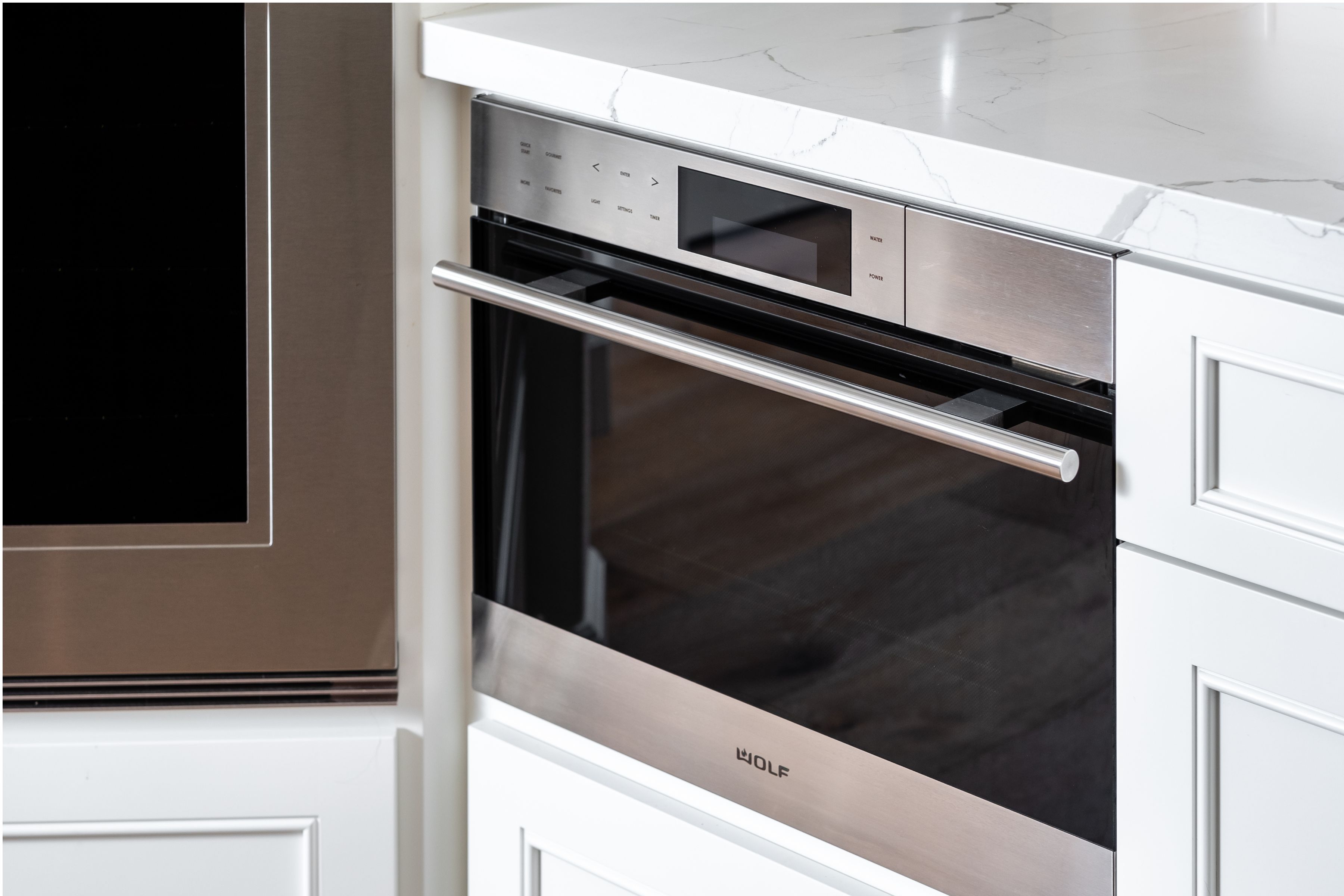 Built-in stainless wall oven set into white kitchen cabinetry beneath a veined quartz counter.