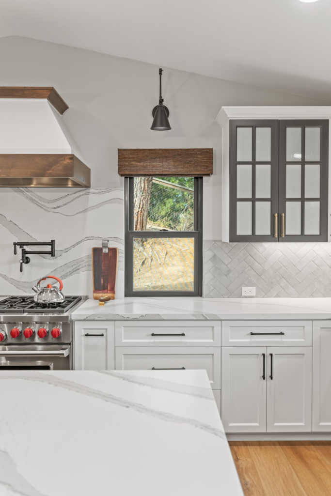 Kitchen cooking wall with white cabinetry, dark glass-front uppers, herringbone tile, pot filler and window.