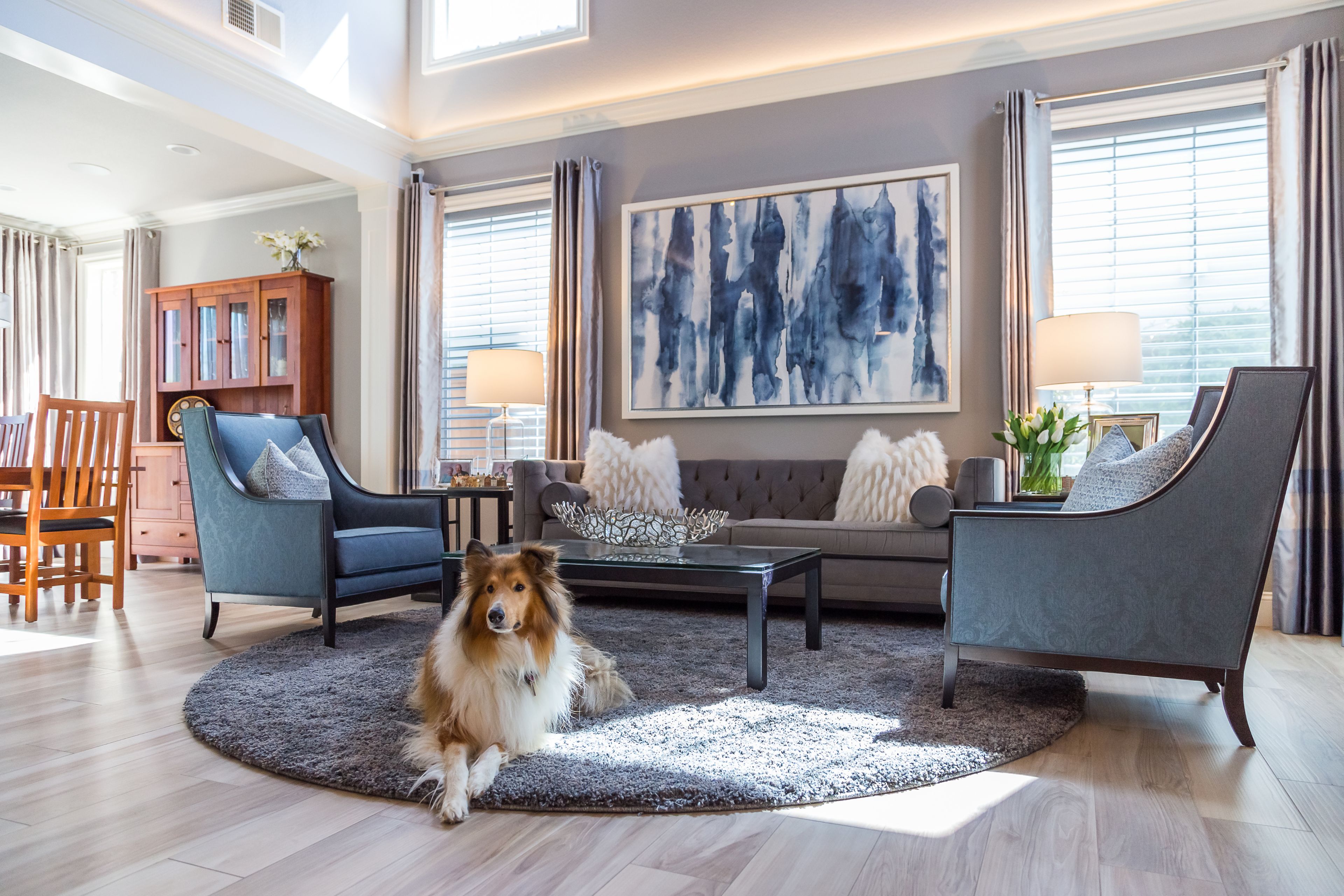Living room with tall windows, gray seating, wood flooring and open connection to the dining area.