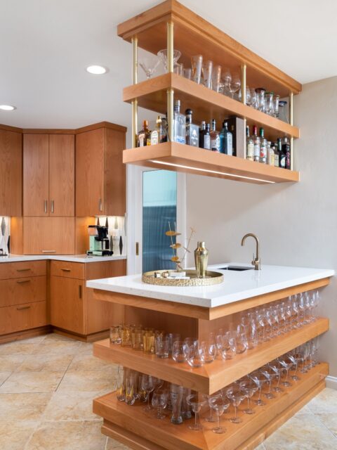 Wet bar with suspended wood shelving, brass supports, white countertop and open glassware storage