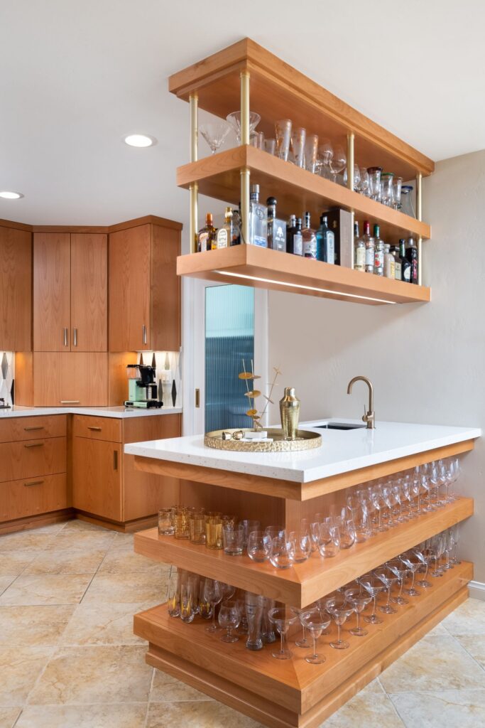 Wet bar with suspended wood shelving, brass supports, white countertop and open glassware storage