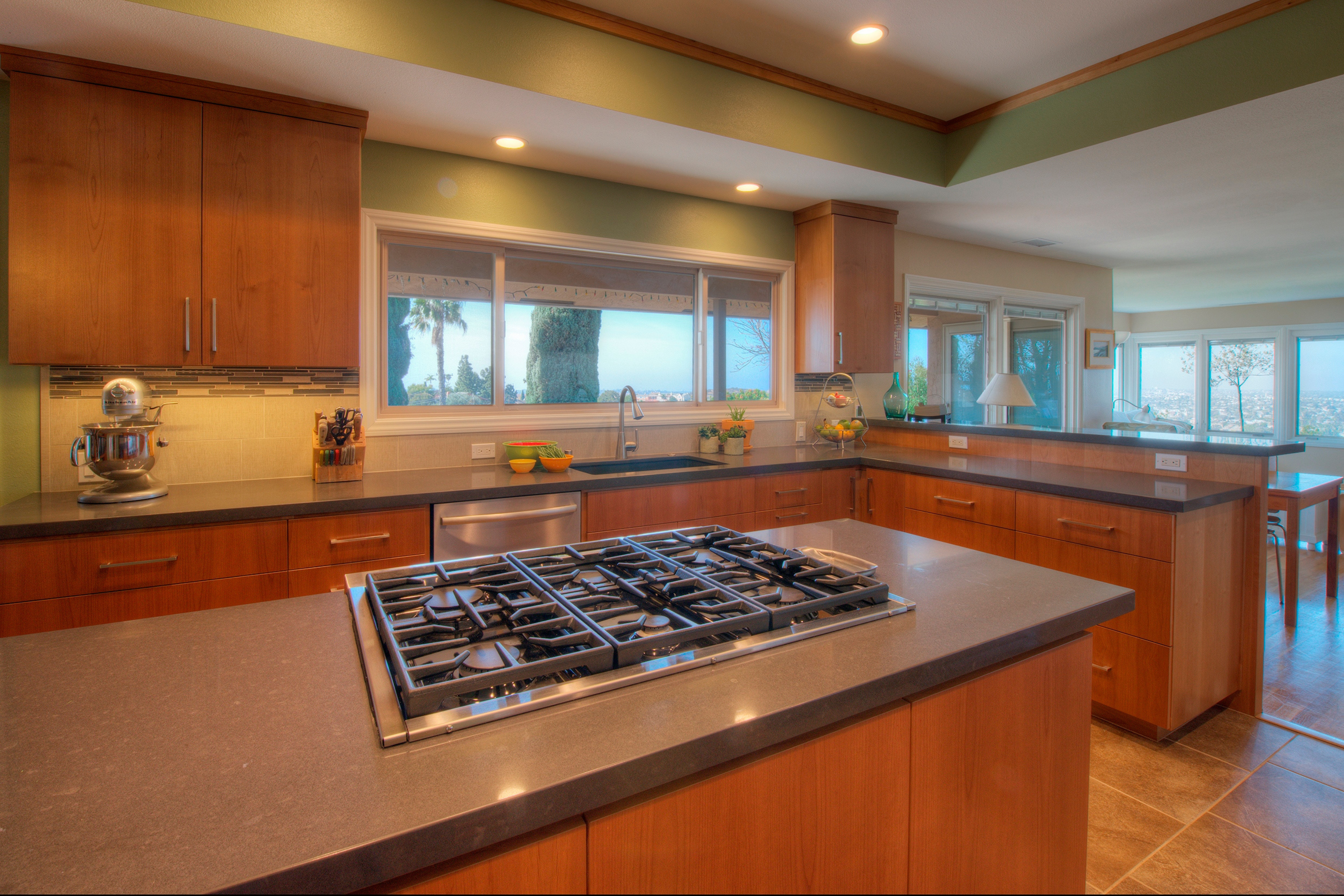Remodeled kitchen with wood cabinets, cooktop island, gray counters, wide sink window and tile flooring.