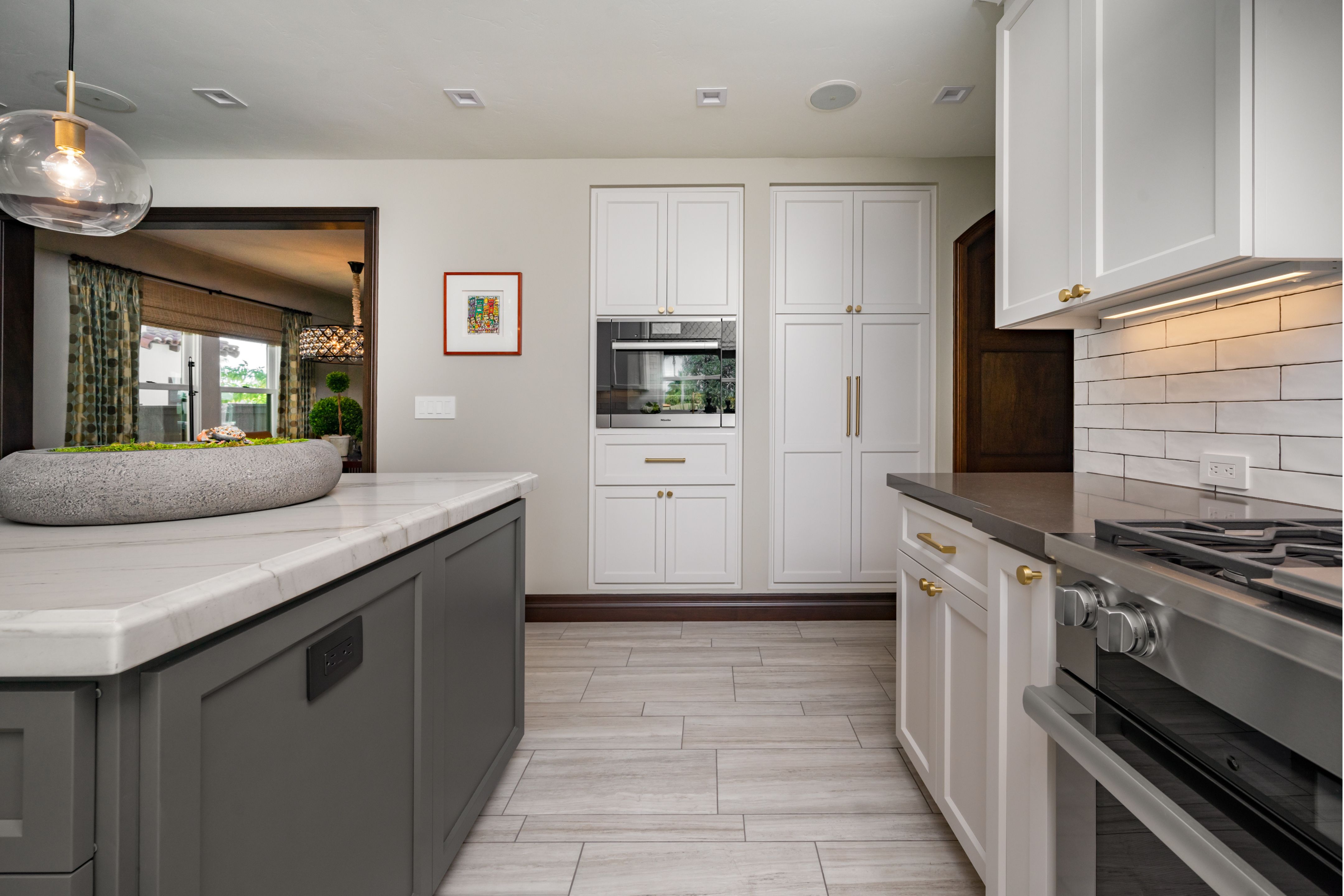 Kitchen work aisle with gray island, white cabinets, built-in oven, brass hardware and light tile flooring