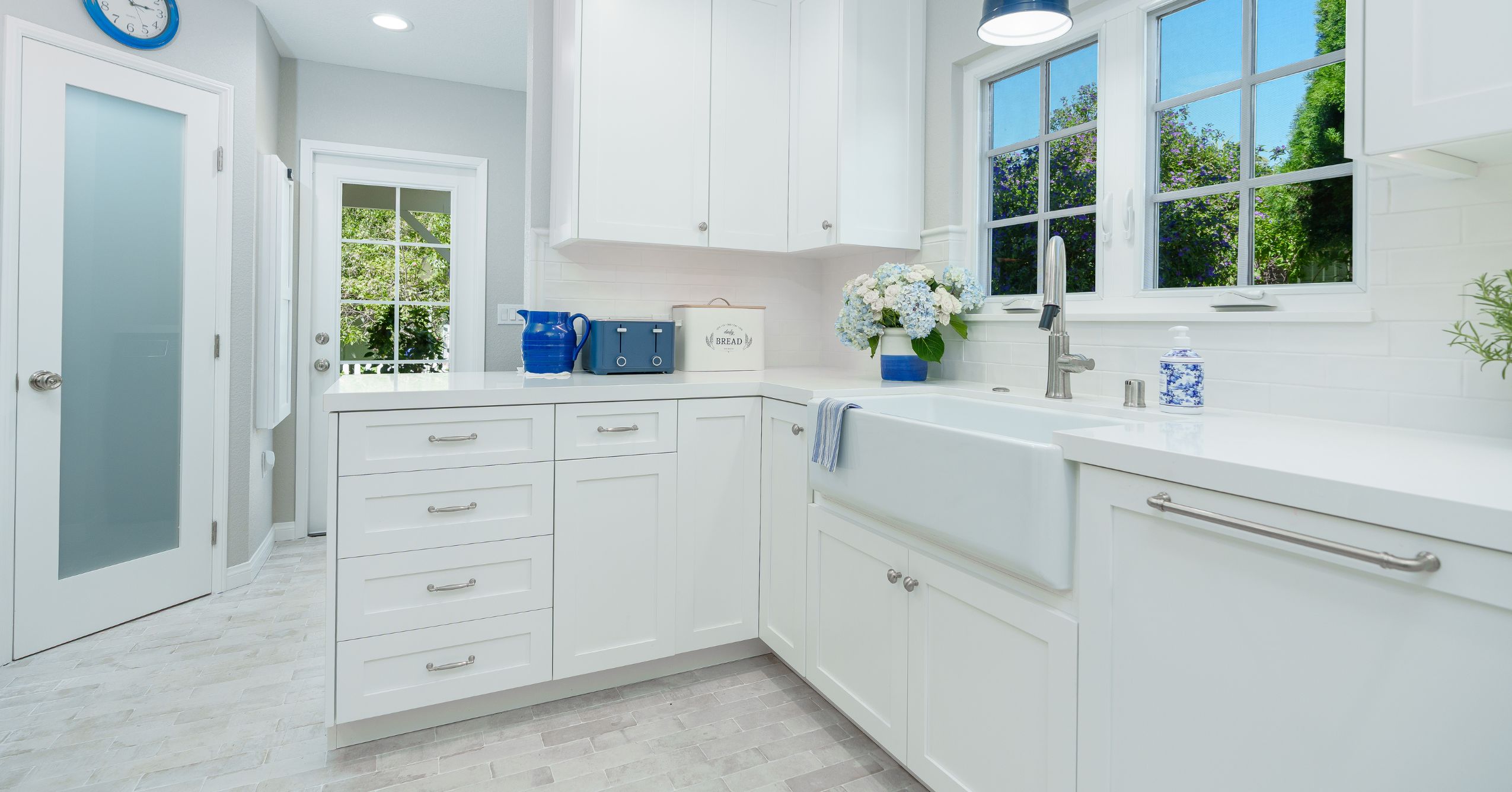 White kitchen with farmhouse sink, divided-light windows, quartz counters, blue pendant, and shaker cabinets.