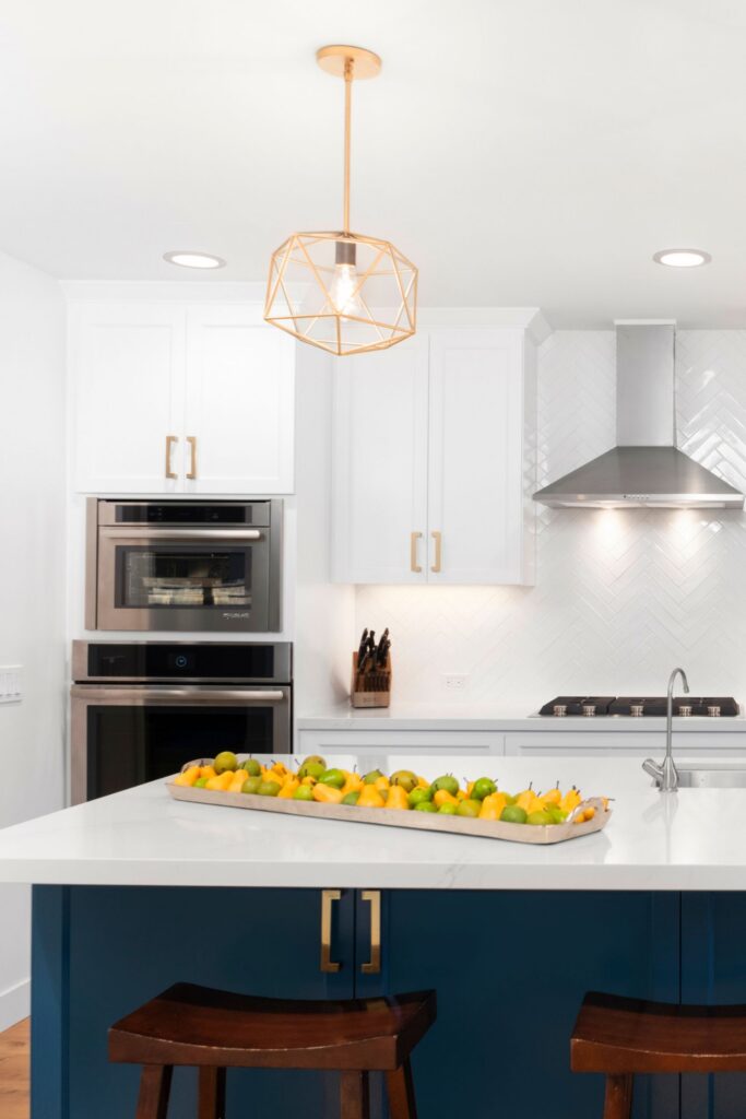 Kitchen with blue island, white countertops, brass geometric pendant lights, white cabinetry, and stainless appliances.