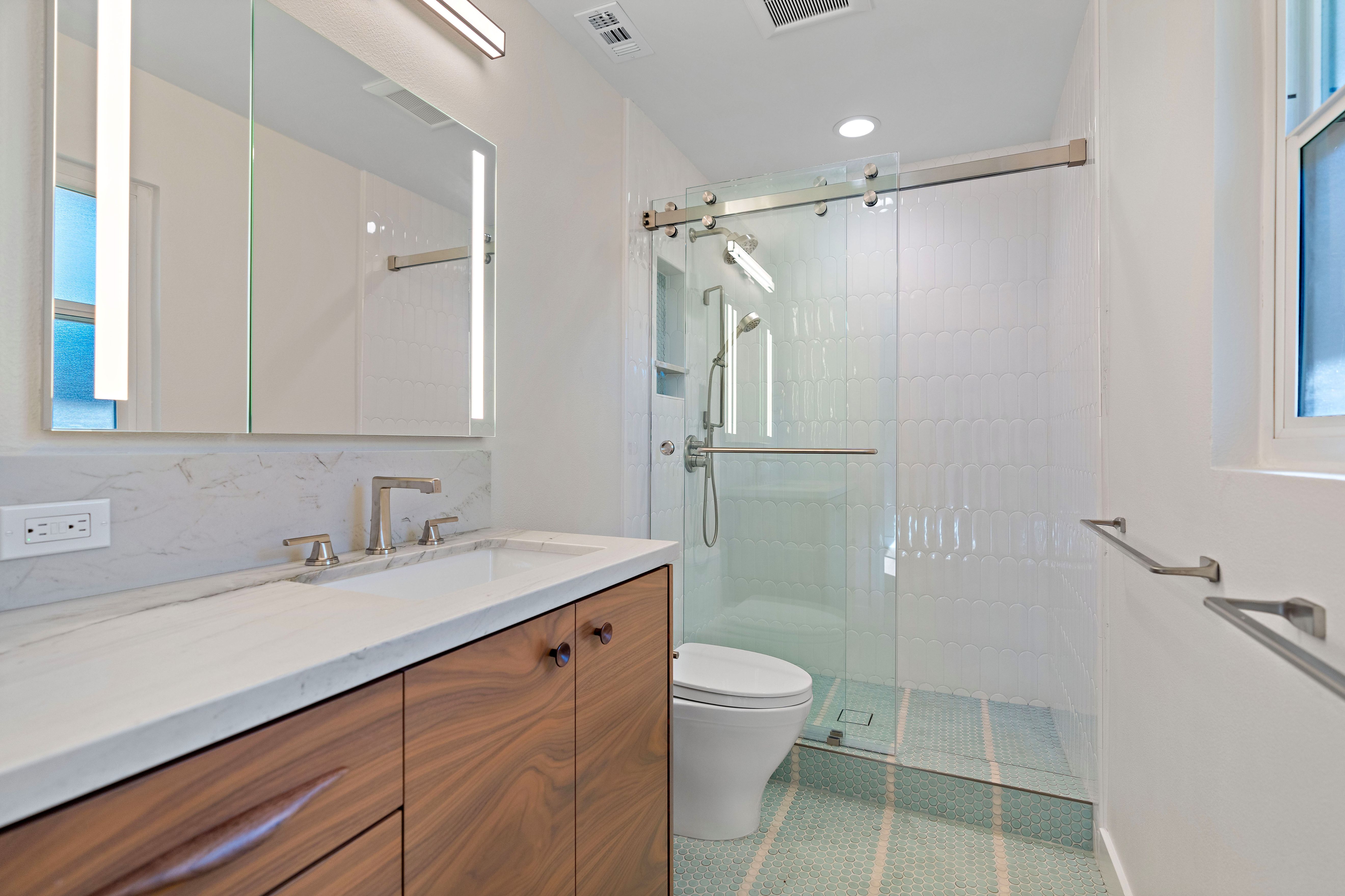Bathroom with walnut vanity, marble-look counter, glass walk-in shower, penny tile floor and illuminated mirror.