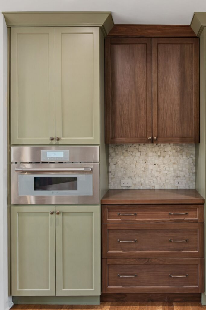 Built-in stainless appliance, sage cabinetry, dark wood drawers, mosaic backsplash, and tall pantry storage.