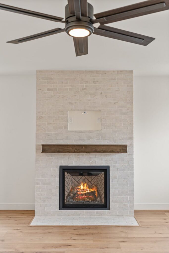 Living room fireplace with pale brick surround, dark wood mantel, built-in firebox and ceiling fan above.