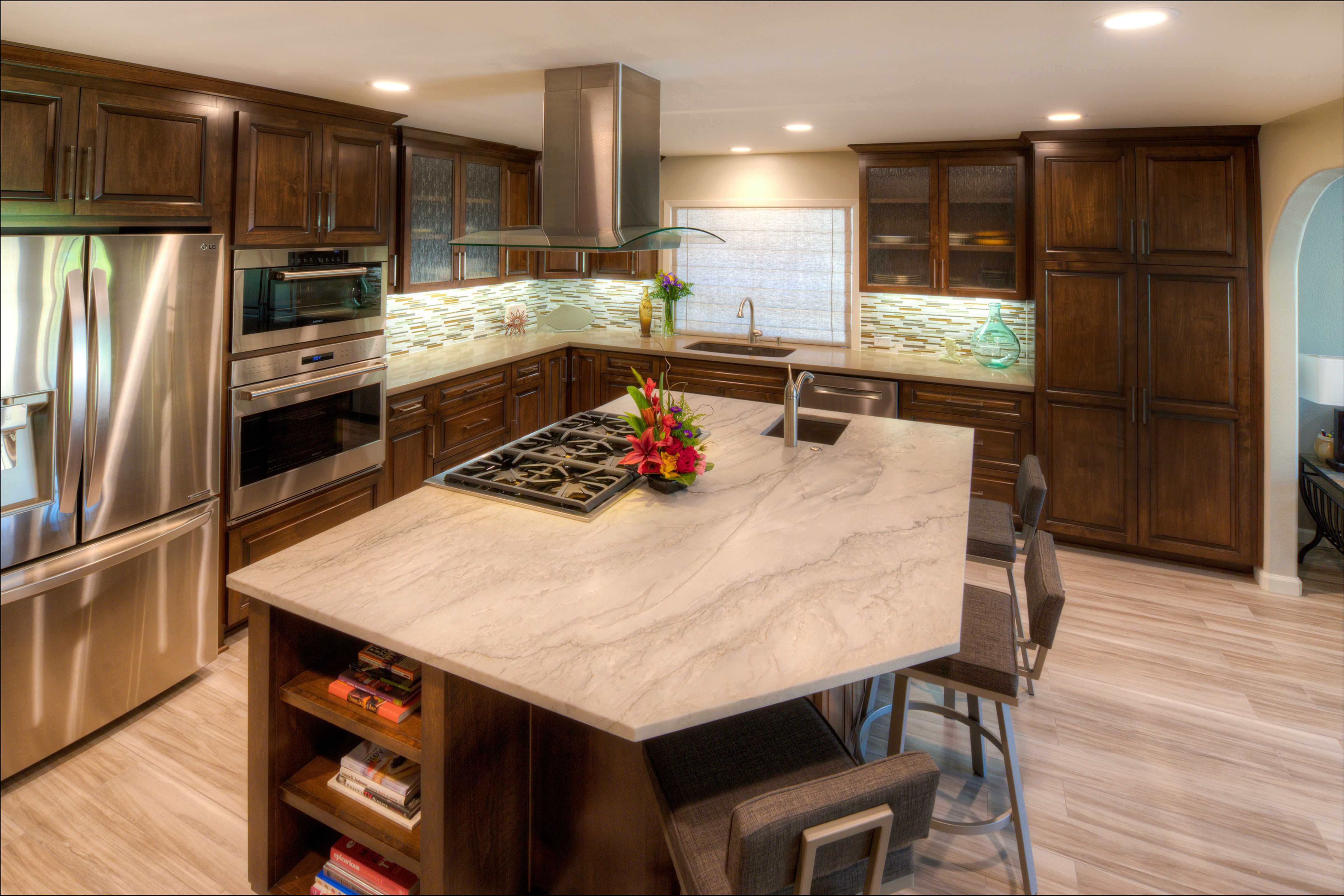 Wood kitchen island with cooktop, prep sink and seating surrounded by dark cabinetry, mosaic tile and stainless appliances.