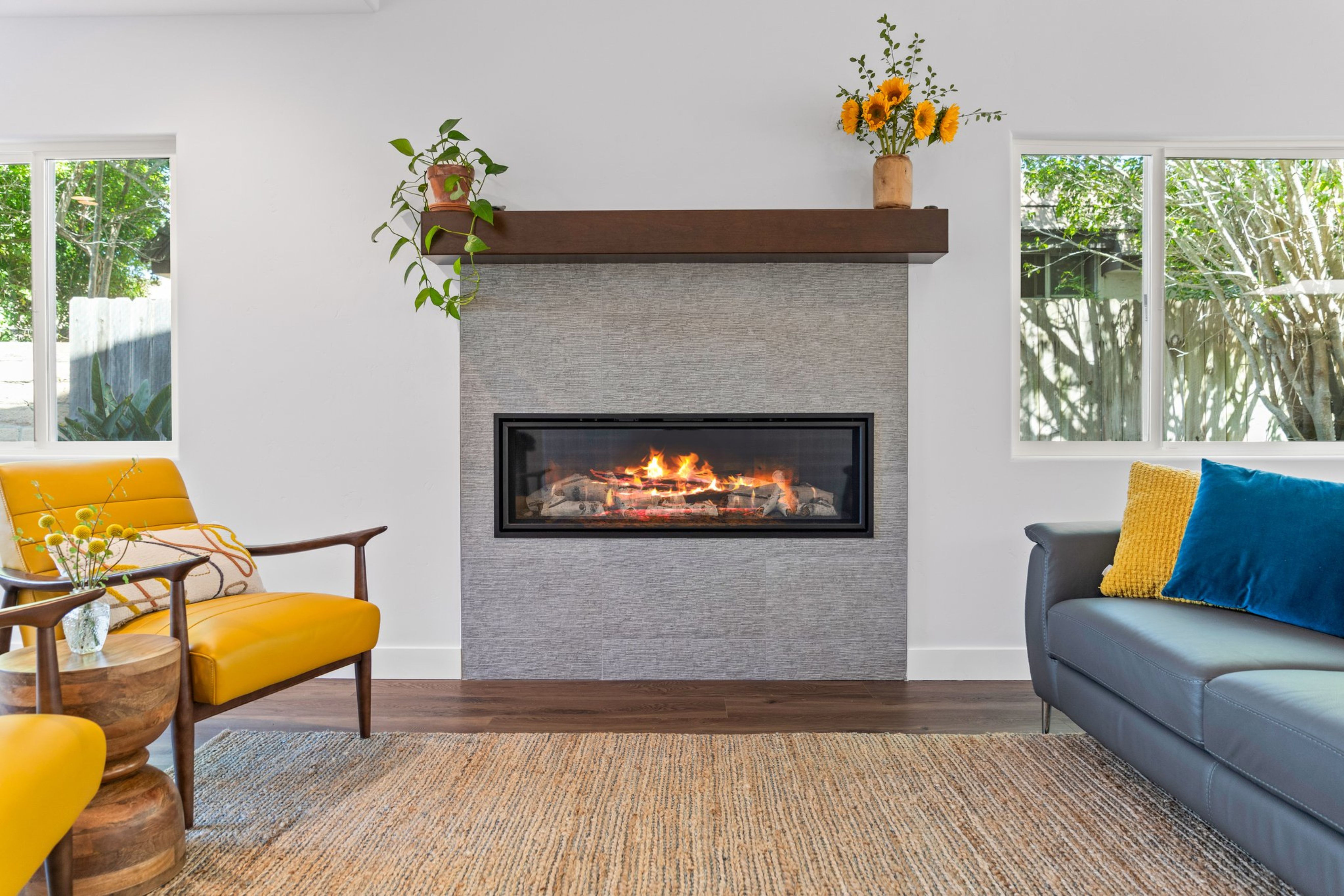 Family room fireplace with gray tile surround, dark wood mantel, large windows and wide-plank flooring.