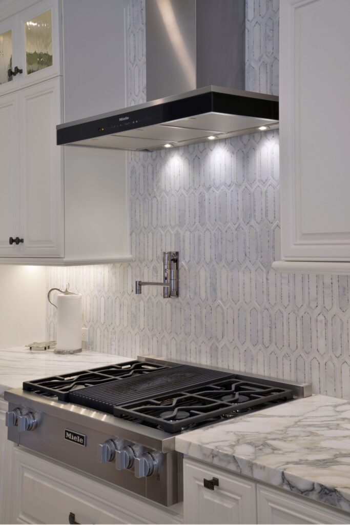 Kitchen cooktop with stainless hood, white cabinetry, marble-look counters and geometric tile backsplash