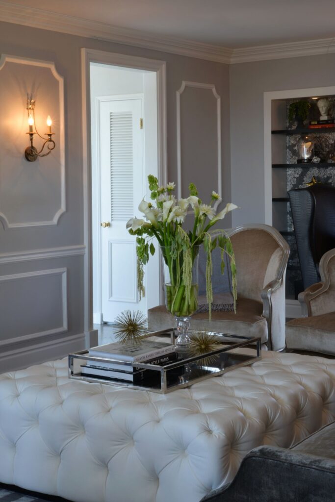 Formal living room with tufted ottoman, gray paneled walls, upholstered seating and wallpapered display niche