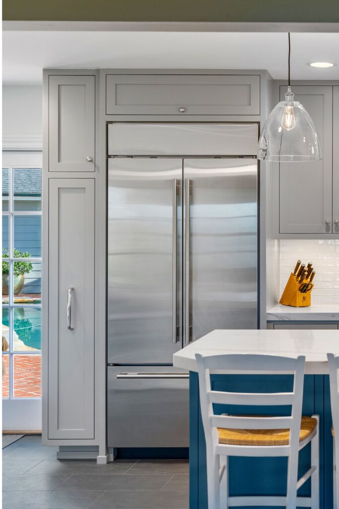 Stainless refrigerator framed by custom gray inset cabinetry beside blue island seating and clear glass pendant lighting.