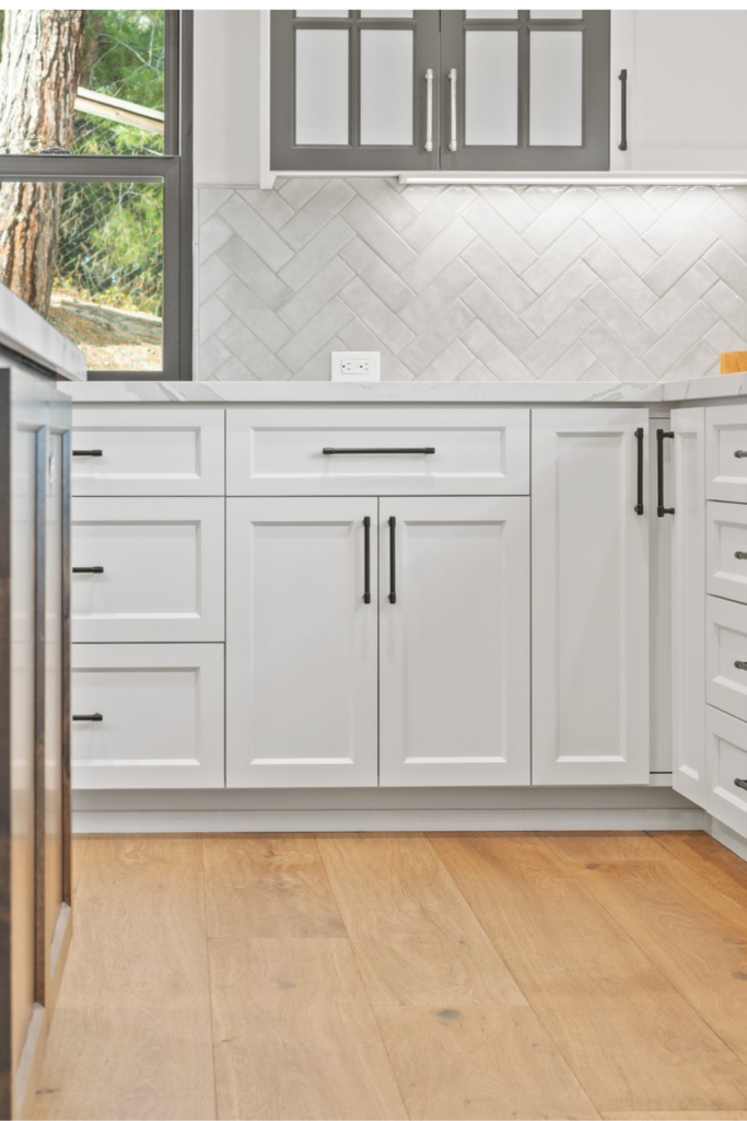 White kitchen base cabinets with black hardware, herringbone backsplash, glass-front uppers and wide-plank wood flooring.