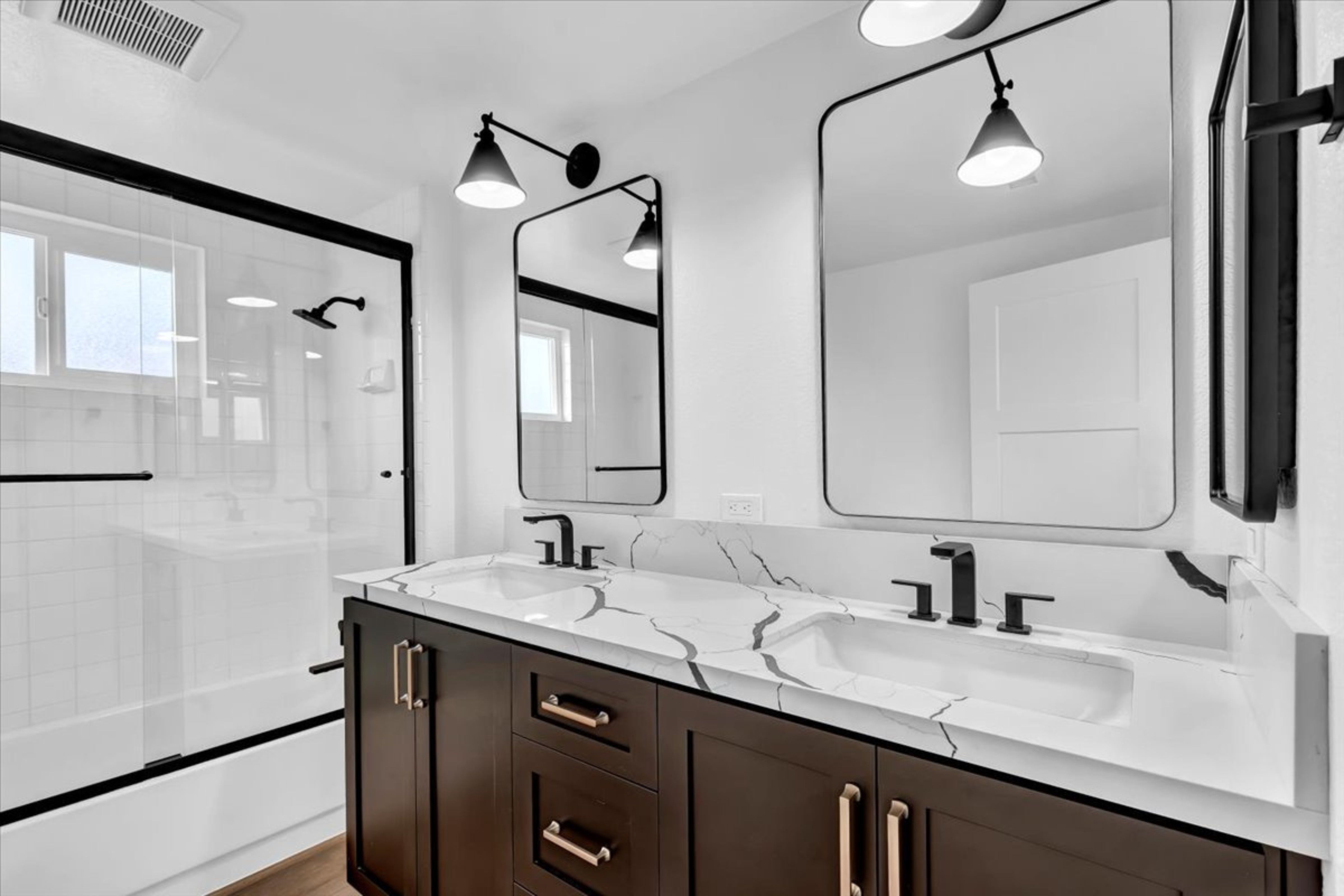Remodeled bathroom with dark double vanity, veined white countertop, black fixtures, mirrors and glass-enclosed shower.
