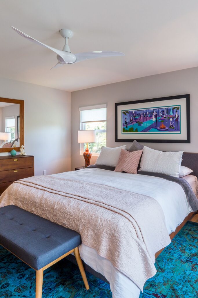 Primary bedroom with white bedding, blue area rug, wood furniture, ceiling fan and framed artwork