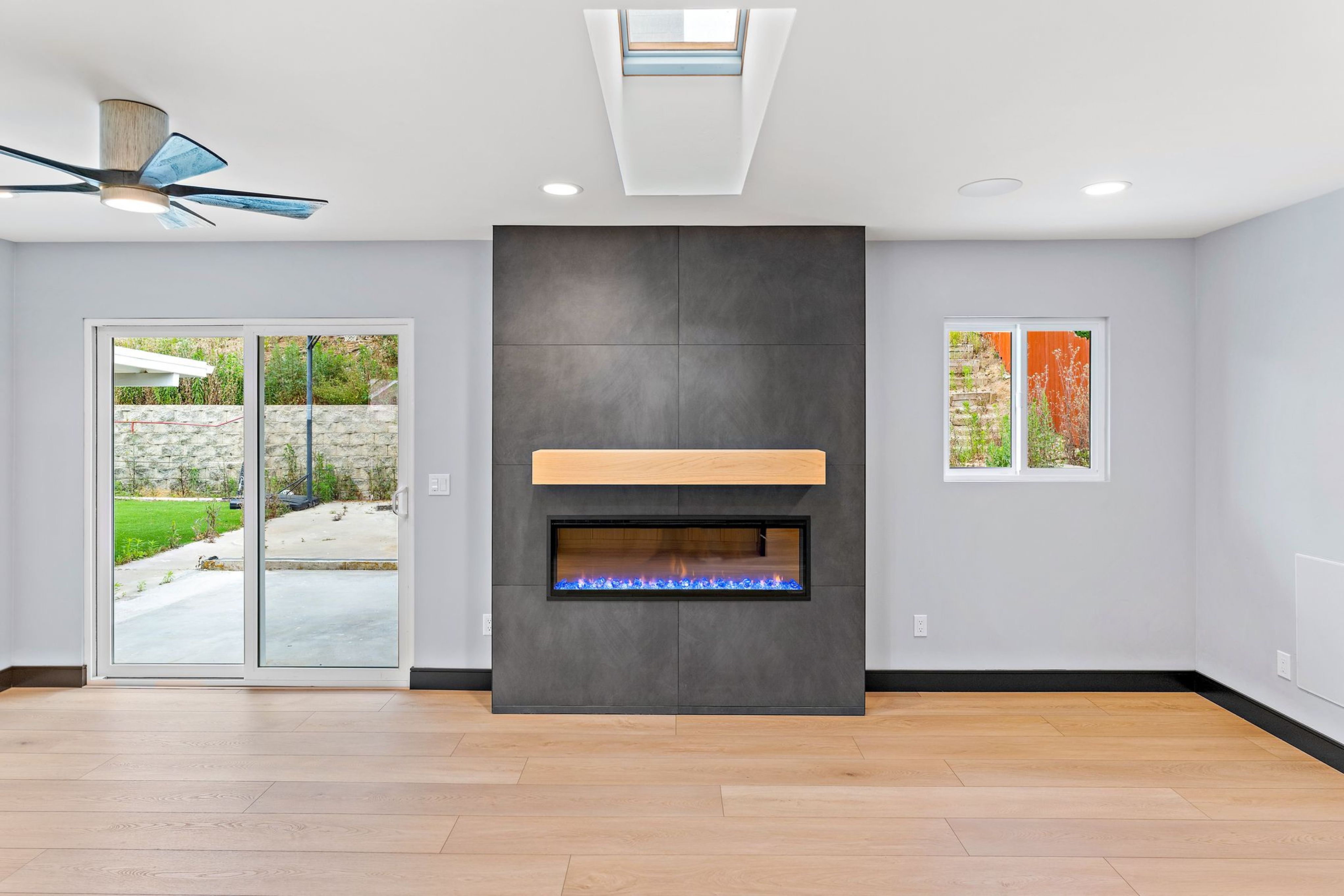 Open living room with dark fireplace surround, wood mantel, skylight, ceiling fan and sliding glass door