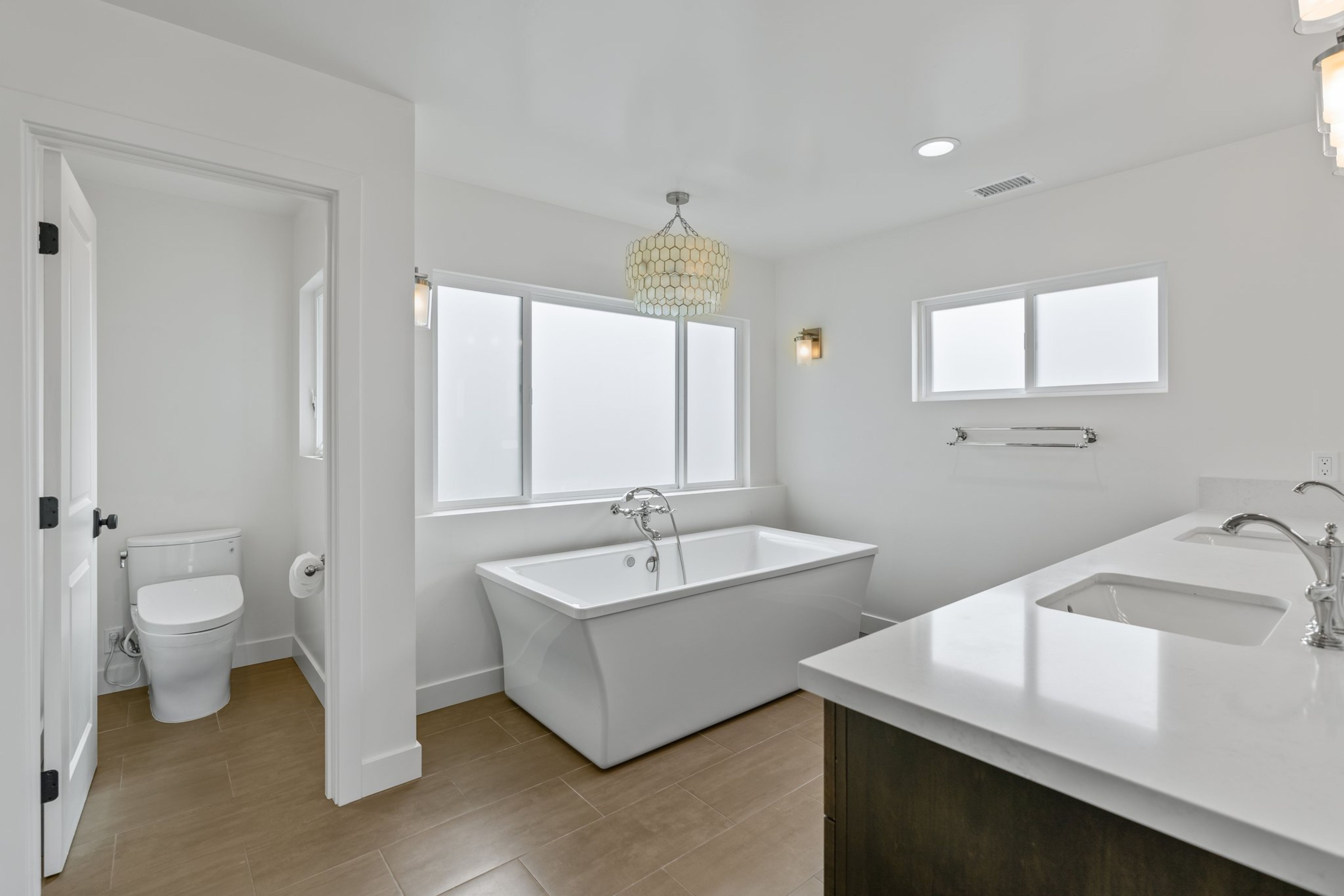 Primary bathroom with freestanding tub, double vanity, separate toilet area, large windows and white countertops.
