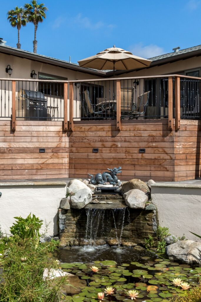 Rear wood deck with metal railing, outdoor dining area and grill overlooking a landscaped pond and waterfall