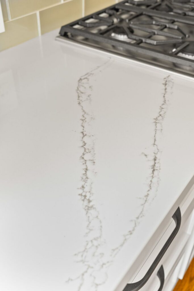 Veined white kitchen countertop beside a gas cooktop with light rectangular tile backsplash.