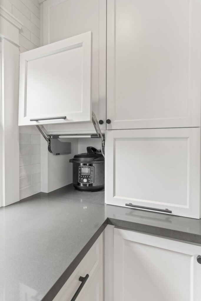 Lift-up white cabinet storing a countertop pressure cooker above gray counters and lower shaker cabinetry.