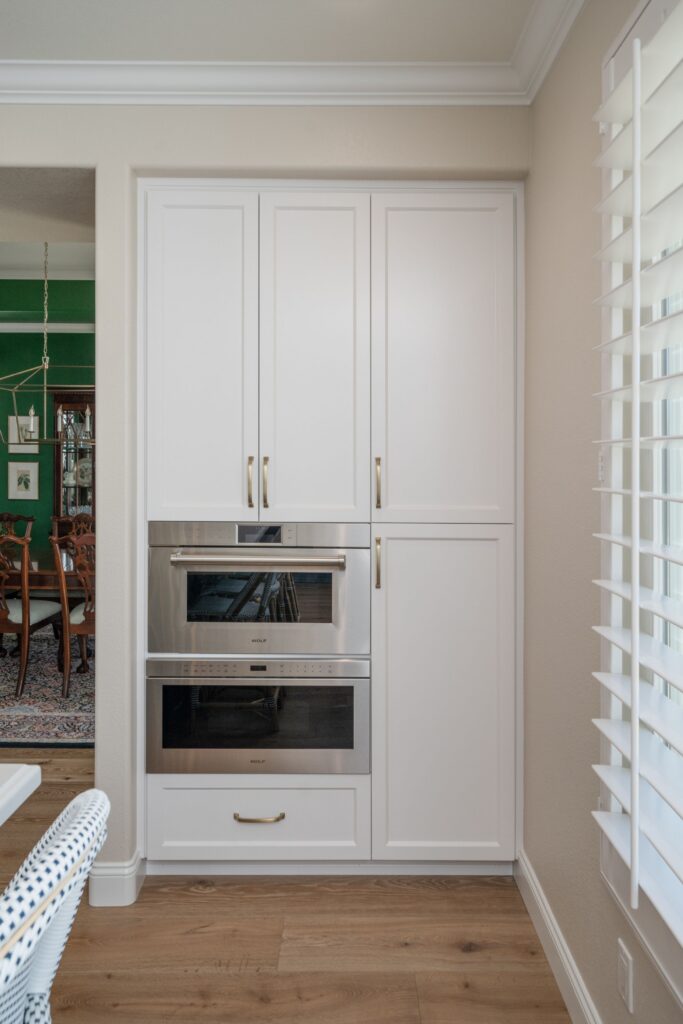 Kitchen appliance wall with white paneled cabinetry, stacked stainless ovens, brass hardware and plantation shutters.