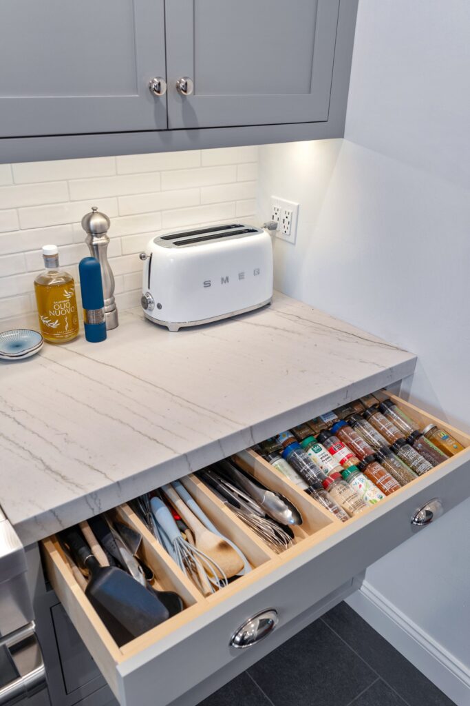Open kitchen drawer with divided utensil and spice storage beneath gray cabinetry and white tile backsplash.