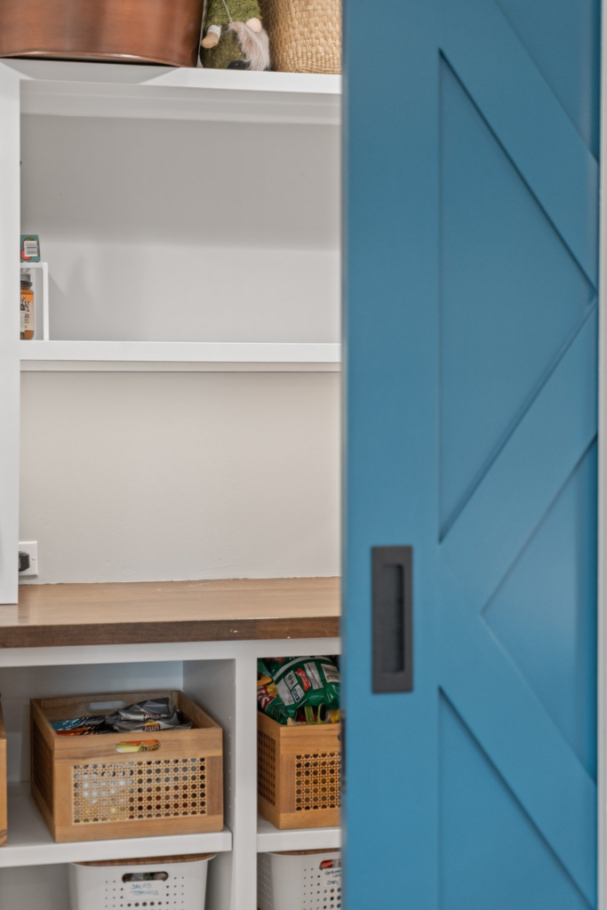 Pantry with blue sliding barn door, white built-in shelving, wood counter and organized storage baskets.