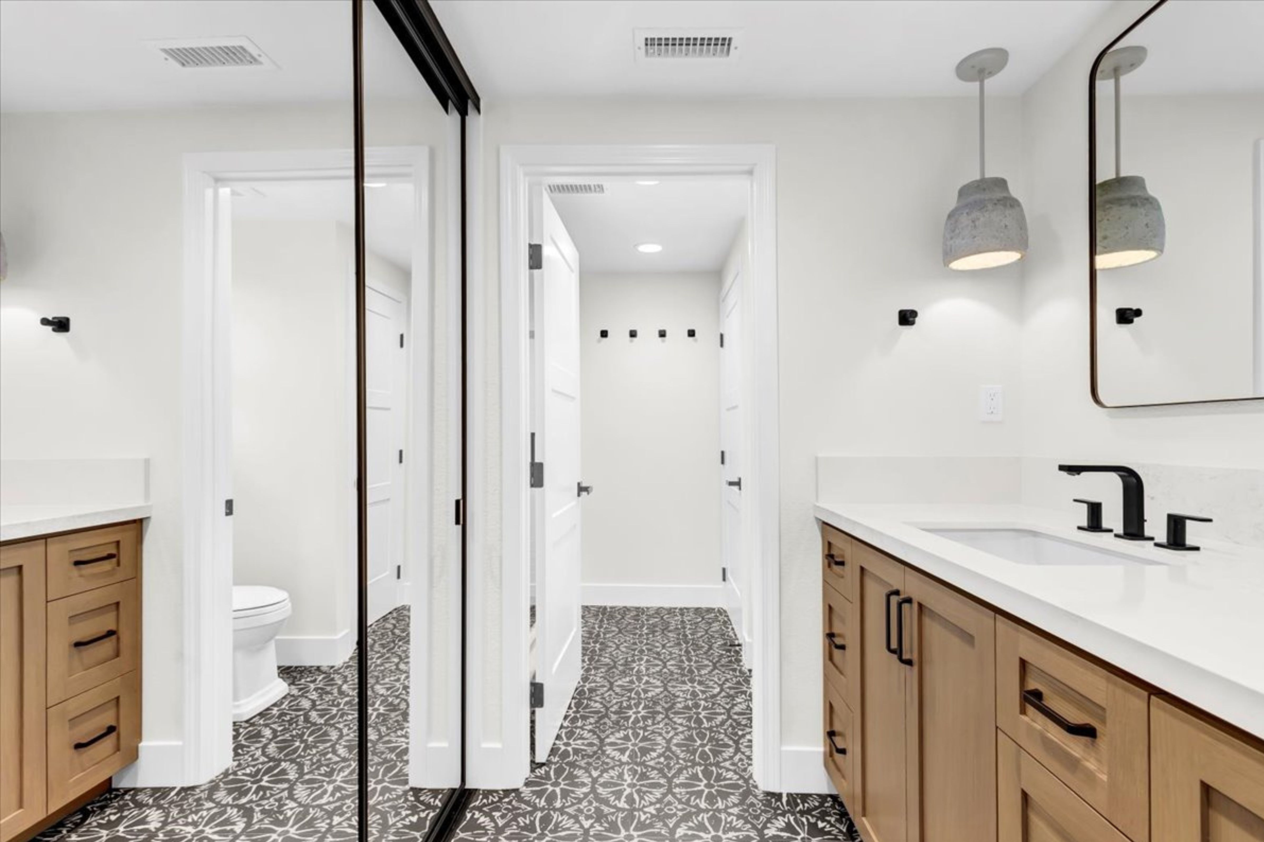 Bathroom vanity with light wood cabinetry, white countertop, patterned floor tile, black fixtures and mirrored closet doors.