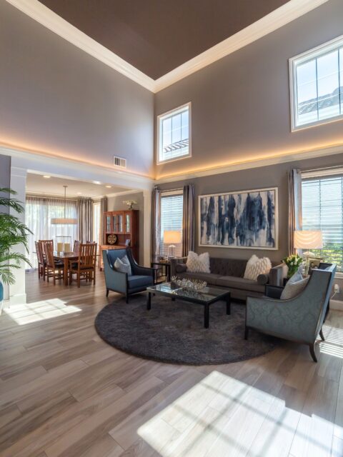 Two-story living room with clerestory windows, cove lighting, gray seating and open connection to the dining room.