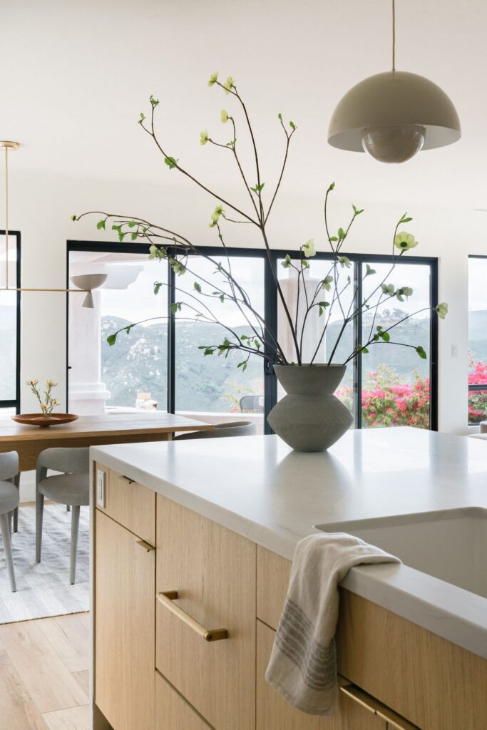 Kitchen island with light wood cabinetry, white countertop, floral arrangement and black-framed doors to the view