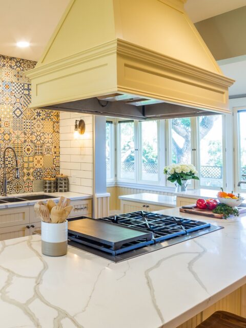 Kitchen island cooktop with veined white countertop, oversized cream hood and patterned tile sink wall