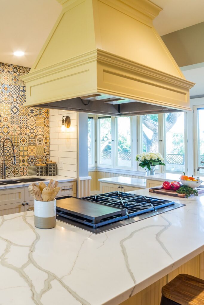 Kitchen island cooktop with veined white countertop, oversized cream hood and patterned tile sink wall